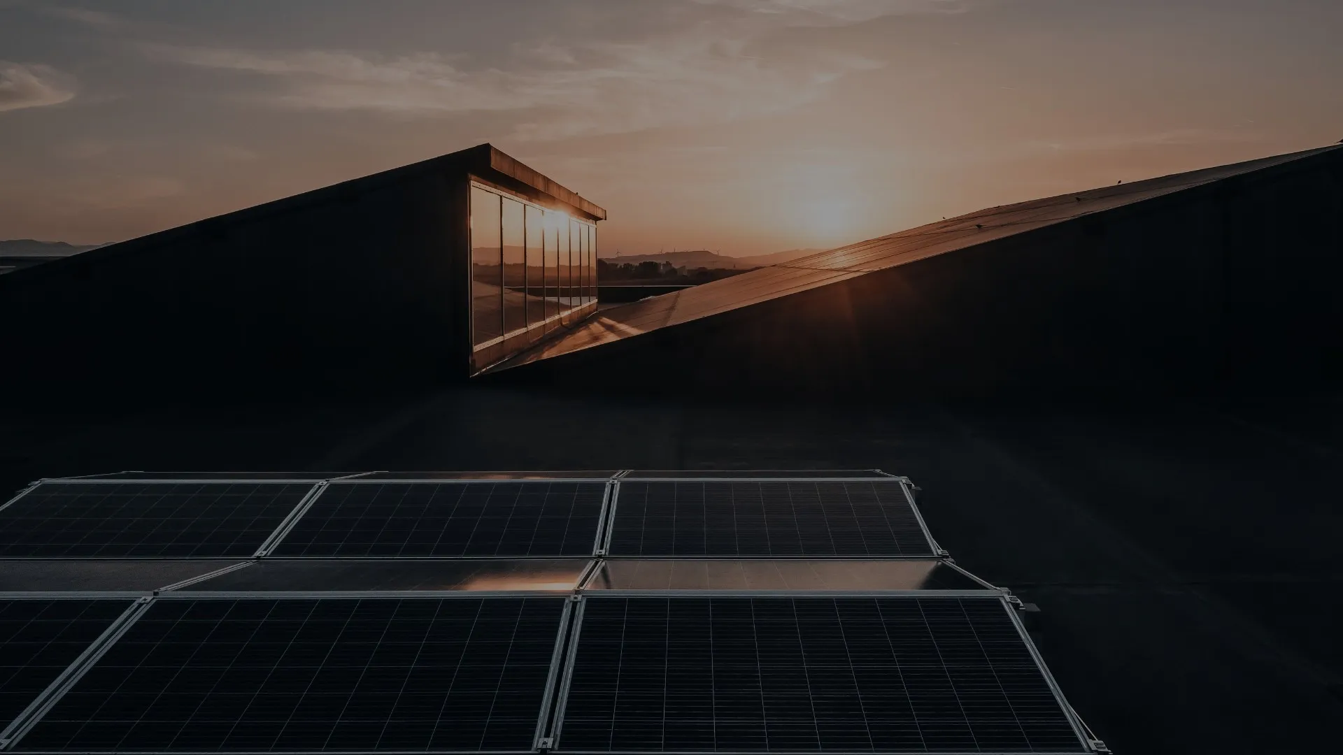 Solarmodule auf einem Flachdach, dahinter ein Gebäude mit Glasfassade im Abendlicht