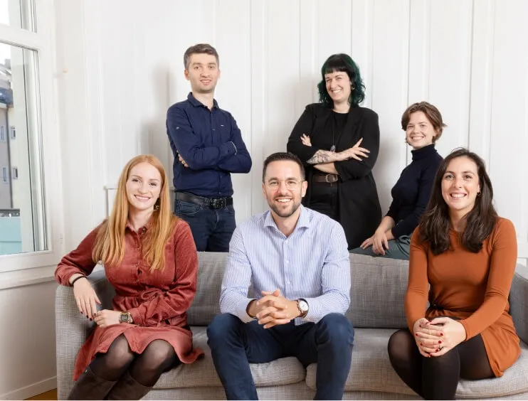 Sechs Mitglieder des Eturnity-Teams lächeln gemeinsam auf und hinter einem grauen Sofa in einem hellen Raum