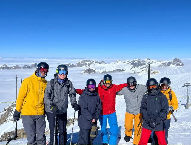 Acht Skifahrende mit Helm und Skibrille auf einem sonnigen Berggipfel über dem Nebelmeer