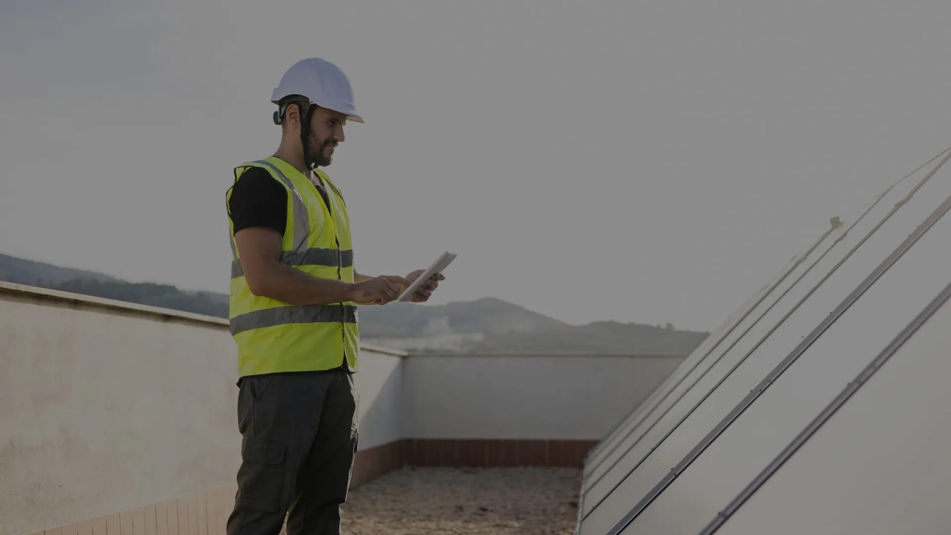 Un installateur en casque et gilet de sécurité consulte sa tablette sur un toit plat, à côté d’une rangée de panneaux solaires