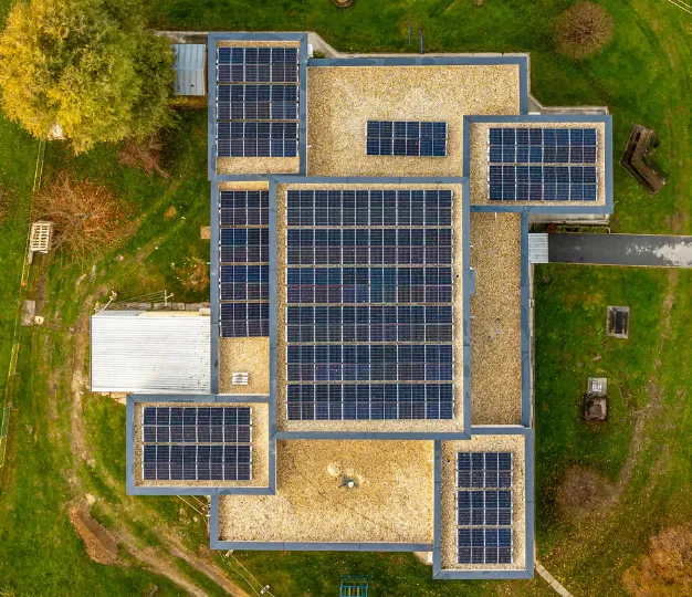 Luftaufnahme eines Flachdachs mit mehreren Solarmodulfeldern auf Kies, ringsum Wiesen