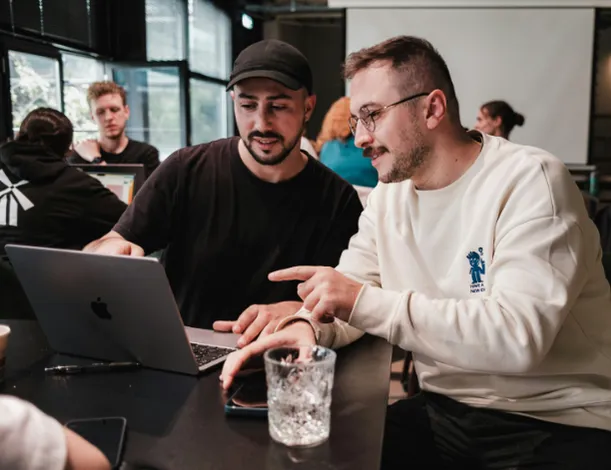 Zwei Männer an einem Café-Tisch schauen auf einen Laptop, einer zeigt auf den Bildschirm